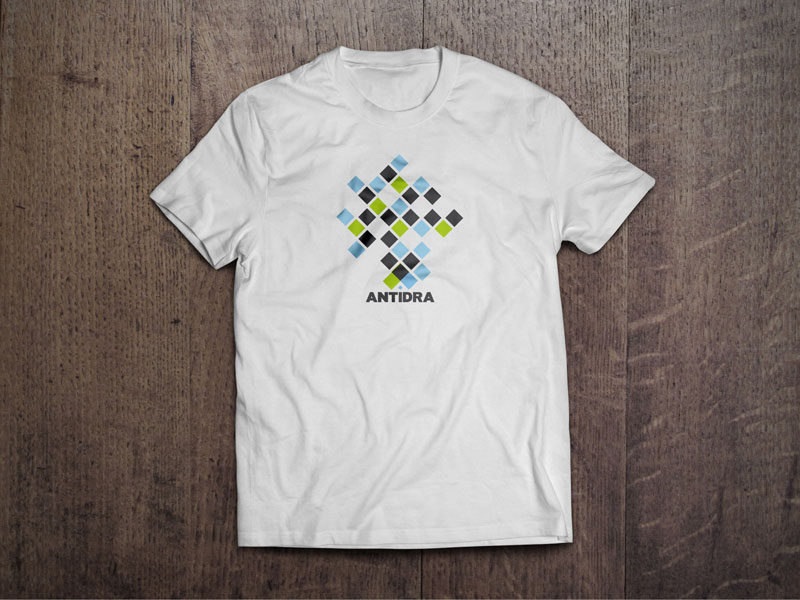T-shirt design for the company Antidra from Croatia. The picture shows a white T-shirt on a wooden base. The T-shirt is printed with a design based on the visual identity of the company Antidra in characteristic green, blue and white squares.