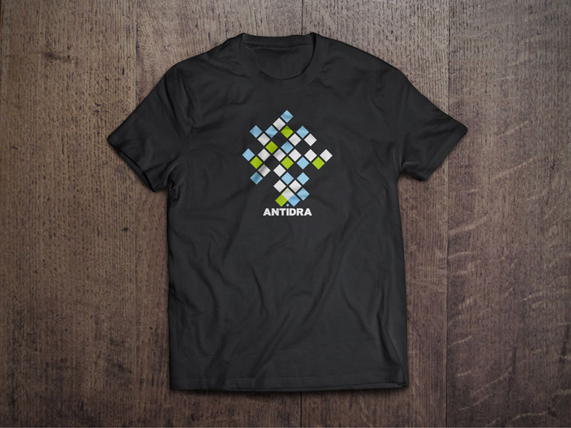 Dizajn T-shirt promotivne majice za tvrtku Antidra iz Hrvatske. Na slici je prikazana crna majica koja se nalazi na drvenoj podlozi. Na majici je otisnut dizajn baziran na vizualnom identitetu tvrtke Antidra u karakterističnim zelenim, plavim i bijelim kvadratićima.
