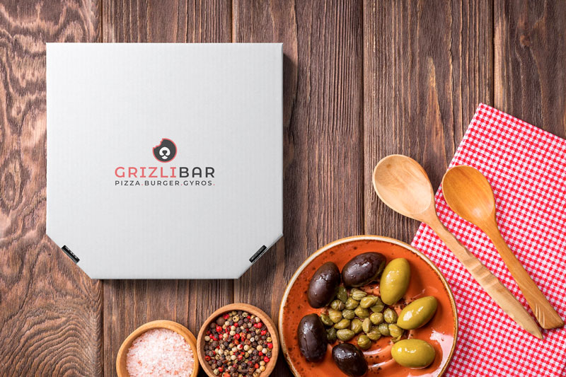 Izrada vizualnog identiteta za restoran Grlizi Bar iz Zagreba, Hrvatska. Na slici je bijela kutija za pizzu na drvenoj površini s logom Grizli Bara. Na stolu su žlice, salvete, masline i začini.