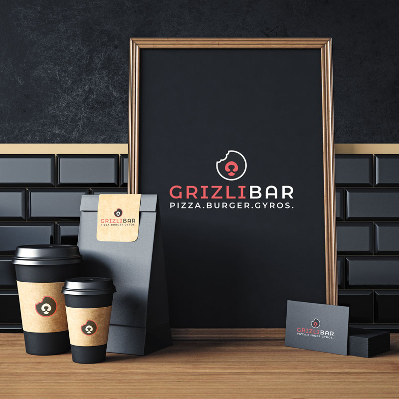 Design of the logo for Grizli Bar in Zagreb. The picture shows a wooden table and a dark gray brick wall in the background. On the table is a poster framed with a wooden frame with the Grizli bar logo on it. The poster is black, the logo is red and white. Along with the poster, there are also coffee cups with the Grizli bar logo, as well as food bags and business cards.