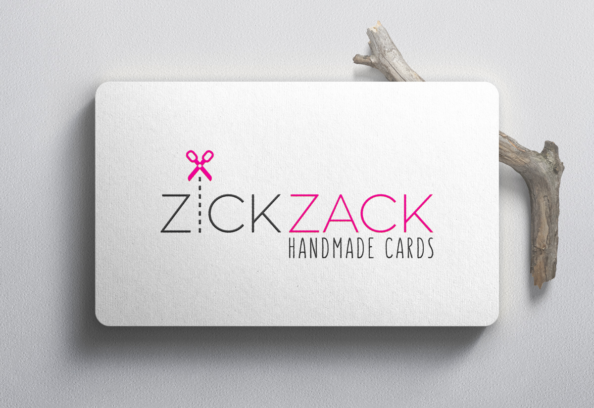Logo design for Zick Zack company from Germany. The picture shows the logo on a business card. The business card is placed on the table and a branch can be seen in the background of one end. The logo is gray pink. Zick is gray and Zack is pink. Instead of the letter I on Zick, there is a broken line, and above it small pink scissors.
