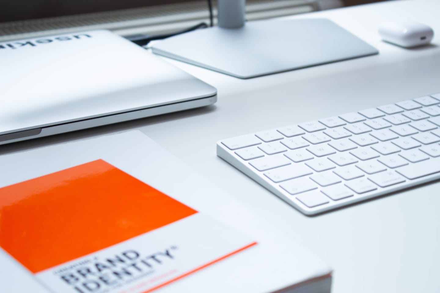 The book “Brand Identity” with an orange cover next to a white keyboard and a laptop on a white desk