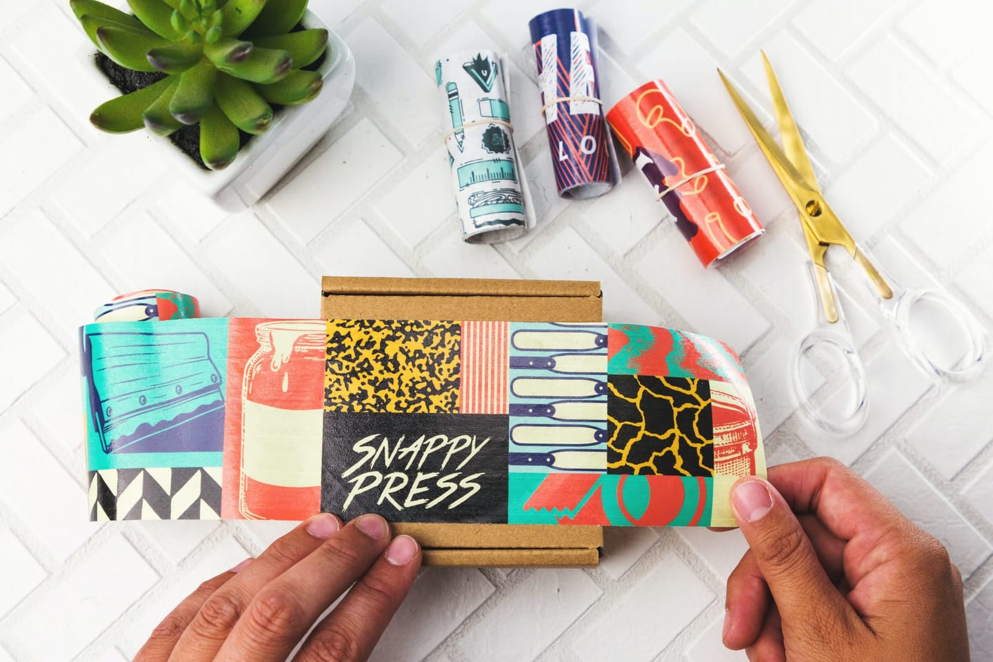 Hands wrapping a cardboard box in a colourful printed band reading “Snappy Press”, next to rolled printed wraps, gold scissors and a succulent