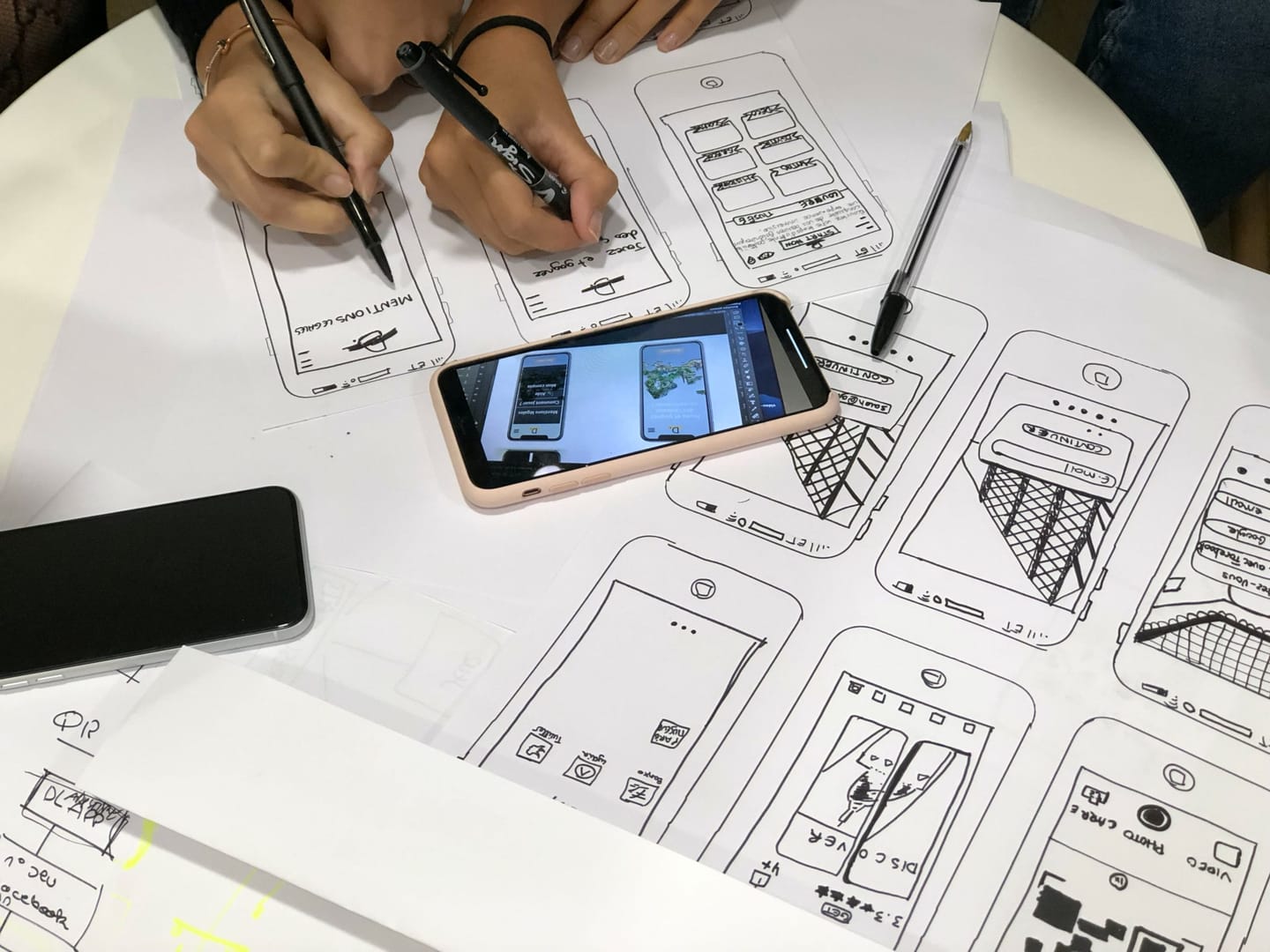 Hand-drawn sketches of mobile app screens on paper, with hands holding markers and two phones on the table