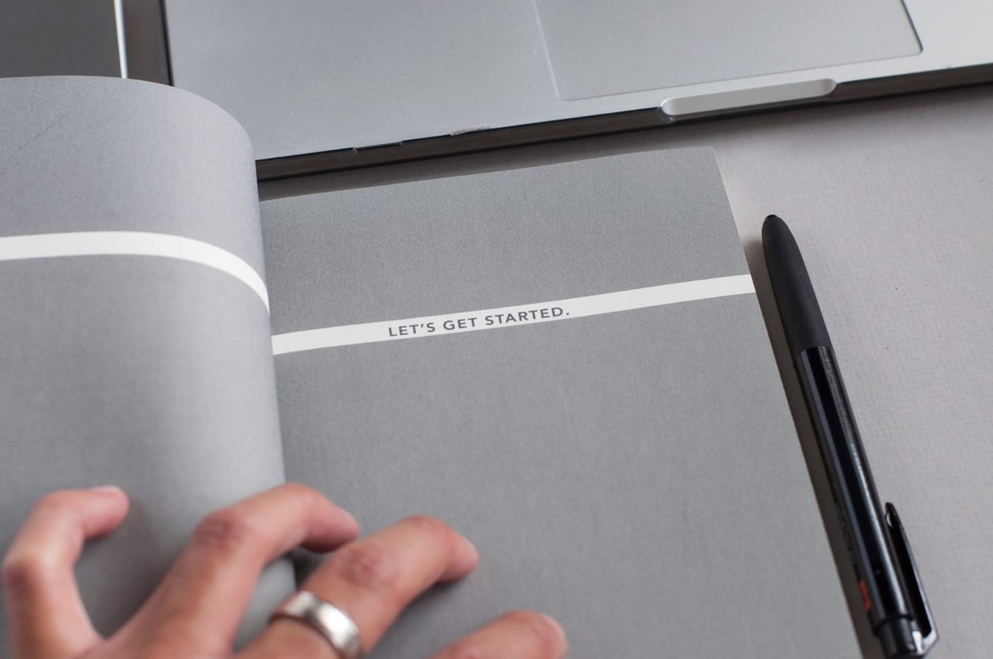 Hand opening a grey booklet with the words “Let’s get started.” next to a laptop and a pen